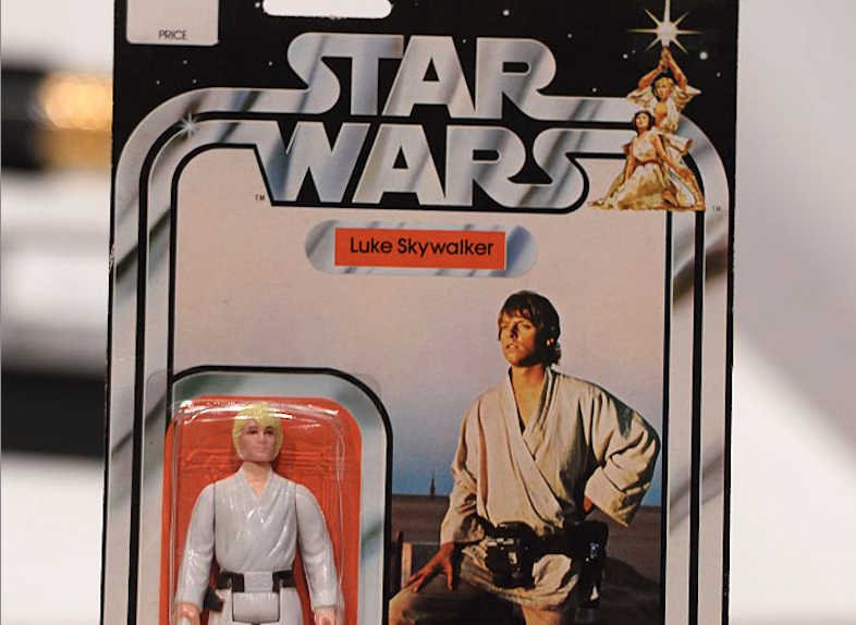 A packaged Star Wars action figure labeled "Luke Skywalker". The packaging features an image of Luke Skywalker in a white outfit with a belt, holding a lightsaber. The figure is displayed within a clear plastic casing.